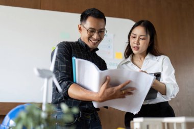 İki mühendis heyecanlı bir şekilde sürdürülebilir bina planlarını tartışıyor, parlak bir ofiste modeller ve planlar sergiliyor..