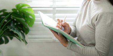 Genç bir kadın ajandasında, bitkilerle dolu canlı bir ev ofisinde organizasyon ve verimliliği vurgulayan notlar yazıyor..