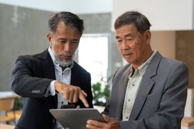 İki kıdemli adam, bir tablet üzerindeki verileri incelerken, modern iş dünyasındaki teknolojinin önemini vurgulayarak görüşlerini paylaşıyorlar..