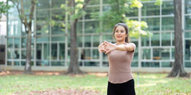 Yeşil bir parkta kol esnetme hareketi yapan bir kadın, dışarıda etkinlik yaparak sağlığını ve uygunluğunu arttırıyor..