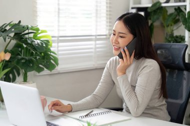 Modern iş hayatı dengesini yansıtan şık bir ev ofisinde dizüstü bilgisayarı üzerinde çalışırken bir telefon görüşmesi sırasında gülümseyen bir kadın..