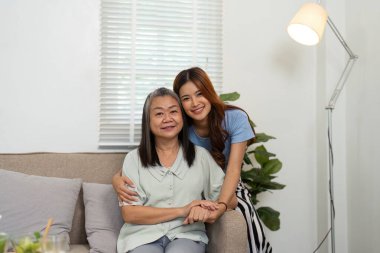 Genç bir kadın ve annesi sevgi dolu bir kucaklaşmayı paylaşıyor, sıcacık bir oturma odasında refahı ve duygusal desteği simgeliyor..