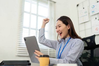 Neşeli, profesyonel bir kadın, parlak ofis alanındaki tabletine bakarken başarısını kutluyor..
