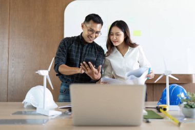 Çevre dostu tasarımları inceleyen ve proje detaylarını tartışan iki mühendis modern bir çalışma alanında akıllı telefon kullanıyor..