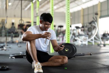 Genç bir adam spor salonunda oturup telefonuna bakarken ve yoğun bir egzersizden sonra suyunu yudumlarken dinleniyor..
