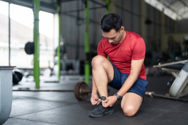 Odaklanmış bir adam spor salonunda diz çöker, spor salonuna başlamadan önce bağcıklarını bağlar. Sağlığın ve hazırlığın vurgulanması.