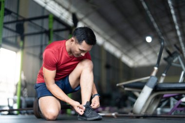 Odaklanmış bir adam spor salonunda diz çöker, spor salonuna başlamadan önce bağcıklarını bağlar. Sağlığın ve hazırlığın vurgulanması.