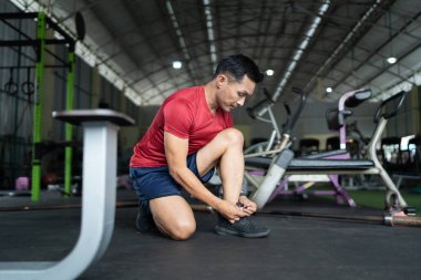 Odaklanmış bir adam spor salonunda diz çöker, spor salonuna başlamadan önce bağcıklarını bağlar. Sağlığın ve hazırlığın vurgulanması.