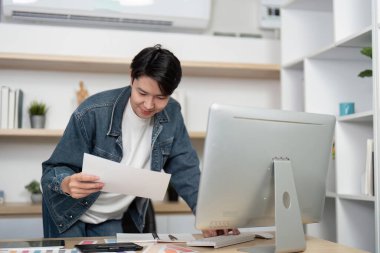 Yazılı materyalleri inceleyen genç bir tasarımcı masasının başında durmuş, tasarım analizinin önemini vurguluyor..