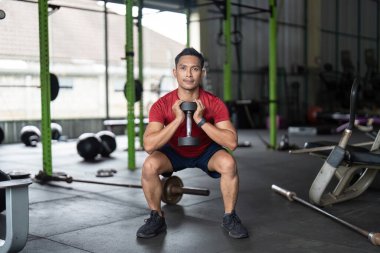Odaklanmış bir adam spor salonunda bir dambılla çömelip, genel sağlık için güç eğitimini ön plana çıkarıyor..
