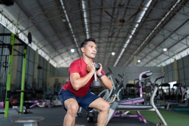 Odaklanmış bir adam bir dambılı tutarken çömelir, spor salonunda vücut gücünü arttırır..