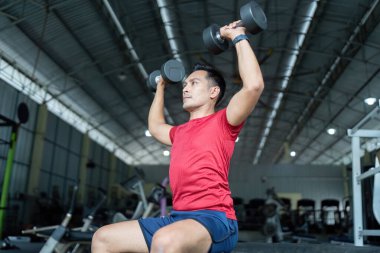 Odaklanmış bir birey, bir fitness ortamında güç eğitimi ve sağlığını ön plana çıkaran bir dambıl presi uygular..