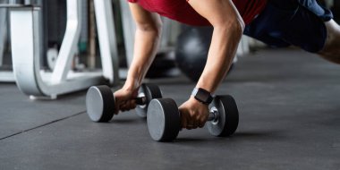 Spor salonunda dambıllar kullanarak şınav çeken bir adam, güç eğitimini ve sağlığını ön plana çıkarır..