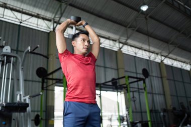 Bir erkek, spor salonundaki güç eğitimini ve sağlığını vurgulayarak, dambıllarla üstten kaldırma gösterisi yapıyor..