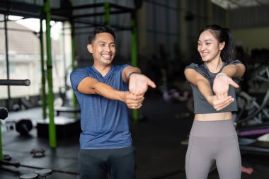 İki arkadaş spor salonunda esneme egzersizleri yapıyor ve antrenmanlarına coşku ve destekle hazırlanıyorlar..