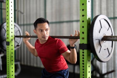 Kendini spora adamış bir erkek sporcu spor salonunda halterle çömelirken şekline odaklanır..