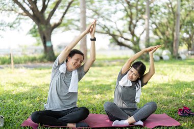 Bir çift, yeşil bir parktaki minder üzerinde yoga yapıyor, paylaşılan aktivitelerle sağlık ve refahı teşvik ediyor..