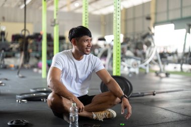Neşeli erkek sporcu spor salonunda oturup egzersiz yaptıktan sonra dinleniyor. Sağlık ve zindelik yaşam tarzını vurgular.