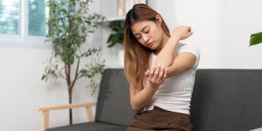 Genç bir kadın, rahat bir ev ortamında rahatsızlık ve acıyı belirterek dirseğini nazikçe ovalıyor. Sağlığa ve sağlığa odaklan..