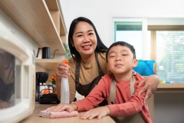 Gülümseyen bir anne temizlik spreyi tutarken oğlu modern bir mutfakta yemek pişirmekten zevk alıyor..