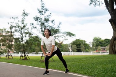 Genç bir kadın, yeşil bir ortamda dinamik esneme ve zindelik hazırlıklarının vurgulanması için açık havada hamleler yapıyor..