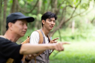 Bir baba ve oğul yürüyüş sırasında bir anı paylaşıyor, yemyeşil ormandaki ilginç manzaralara işaret ederek, bağlarını vurguluyorlar..