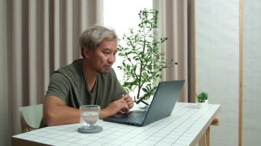 Evde oturan Asyalı bir adam, kendisine verilen bir projenin bütçesini özetlemeye odaklanmıştı. Uzaktan çalışma, sorumluluk ve konsantrasyon gösteriyordu..