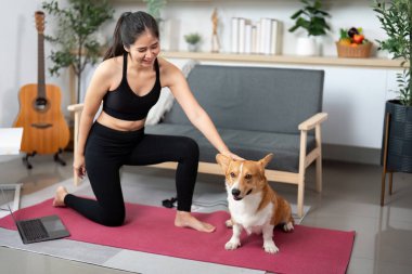 Corgi köpeği yanında otururken bir kadın yoga pozunda diz çöker. Fitness ve evcil hayvan arkadaşlığının uyumunu gösterir..