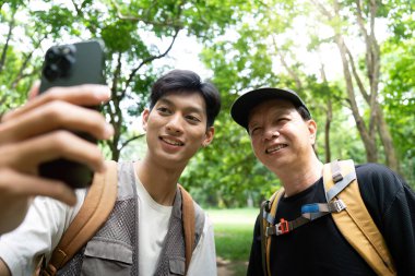 Bir baba ve oğul açık havada eğlenceli bir anı paylaşıyor, yemyeşil bir ortamda selfie çekiyor, bağlarını ve doğaya olan sevgilerini sergiliyorlar..