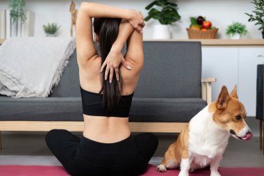Bir kadın, Corgisiyle birlikte yoga minderinde otururken kollarını uzatıyor. Zindelik, sağlık ve duygusal bağlantıyı vurgula.