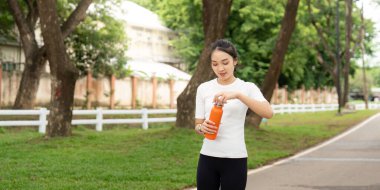 Genç bir kadın parkta yürürken portakal suyu şişesinden serinletici bir içecek içiyor..