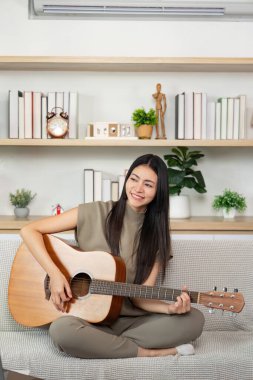 Genç bir kadın rahat bir oturma odasında gitar çalarken gülümsüyor yaratıcılığı ve rahatlığı somutlaştırıyor. Müzik ve neşe kavramı.