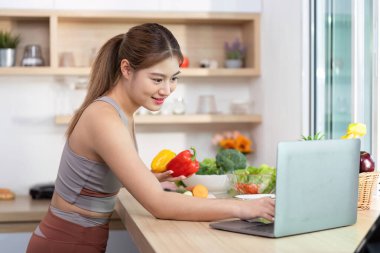 Bir kadın dizüstü bilgisayarla uğraşırken elinde dolmalık biber tutuyor. Parlak bir mutfakta sağlıklı yemek ve sağlığa odaklanıyor..