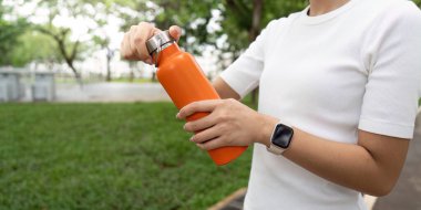 Parktaki genç bir kadın turuncu su şişesinin vidalarını söküyor, idmandan sonra susuz kalmaya hazır..