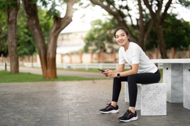 Parktaki bankta mola veren bir kadın telefonunu kontrol ederken gülümsüyor. Doğal bir ortamda spor ve eğlence dengesini temsil eder..