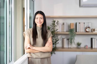 Kendine güveni tam bir kadın pencerenin kenarında durur, şık ev ortamında sıcak bir şekilde gülümser..