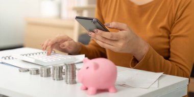 Bir kadın, hesaplarını akıllı bir telefonla yönetirken pembe bir kumbarayı görünür hale getiriyor, dijital finans yönetimini sergiliyor..