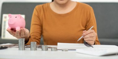 Genç bir kadın aktif olarak mali işlerini yürütüyor, elinde bir kumbara tutuyor ve notlar yazıyor, finansal yetkilendirmeyi ve bütçeyi sembolize ediyor.