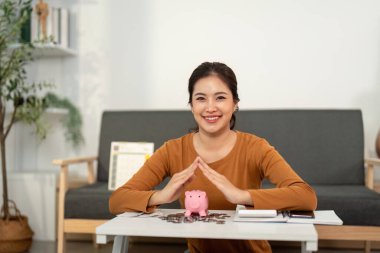 Genç bir kadın, bir kumbarayla otururken güven içinde gülümsüyor, mali güveni ve birikimleri simgeliyor..