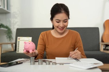 Bütçeyle meşgul gülümseyen genç bir kadın, elinde bir kumbara, defterine yazı yazıyor, mali sorumluluk gösteriyor..