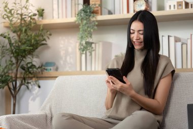 Genç bir kadın akıllı telefonunu rahat bir koltukta kullanırken gülümsüyor. Modern dijital bağlılık ve rahatlamayı yansıtıyor..