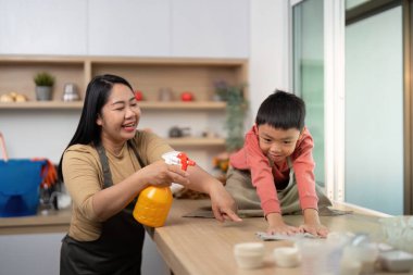 Bir anne temizlik spreyi sıkarken oğlu mutfakta şakacı bir şekilde asistanlık yapıyor. Aile birlikteliğini ve paylaşılan sorumlulukları vurgulamak.