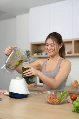 Neşeli bir kadın besleyici bir içecek için taze sebzeleri karıştırır. Parlak bir mutfakta sağlıklı beslenmeyi ve sağlığı teşvik eder..