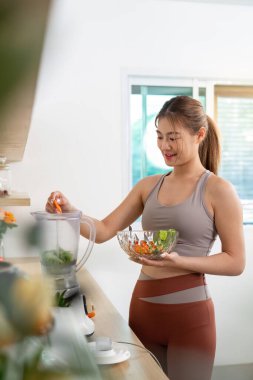 Genç bir kadın, sağlığını ve sağlığını vurgulamak için mutfağında taze sebzelerle smoothie hazırlıyor..