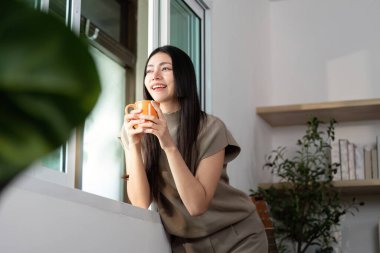 Bir kadın elinde kahve fincanı tutarken gülümsüyor, pencereden dışarı bakıyor, evinde bir neşe ve rahatlama anını somutlaştırıyor.