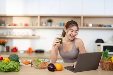 Gülümseyen bir kadın laptopuyla çalışırken yeşil bir smoothie içer. Beslenme ve üretkenliğin karışımını vurgular..