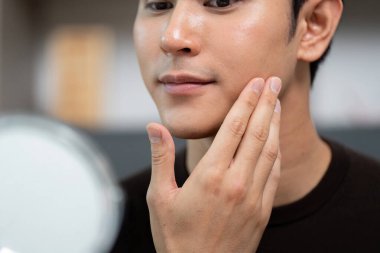 Genç bir adam aynada cildini incelerken nazikçe yüzüne dokunuyor, cilt bakımı ve cilt bakımına odaklanıyor..