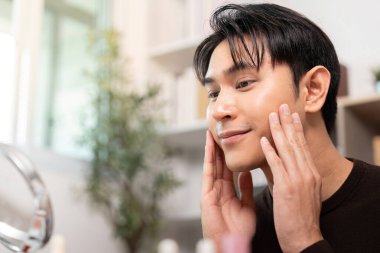 Genç bir adam nazikçe yanaklarına krem sürüyor, modern bir cilt bakım rutini sergiliyor..