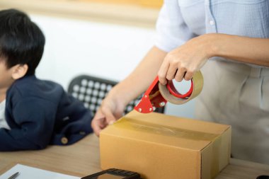 Kendini işine adamış bir anne, çocuğu yanında otururken bir paketi mühürlemek için bantla aile ve internet işinin karışımını resmediyor..