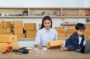 Bir anne, çocuğu yakınlarda oynarken çevrimiçi işi için notlar yazar. Ev ortamındaki iş ve ailenin karışımını betimler..